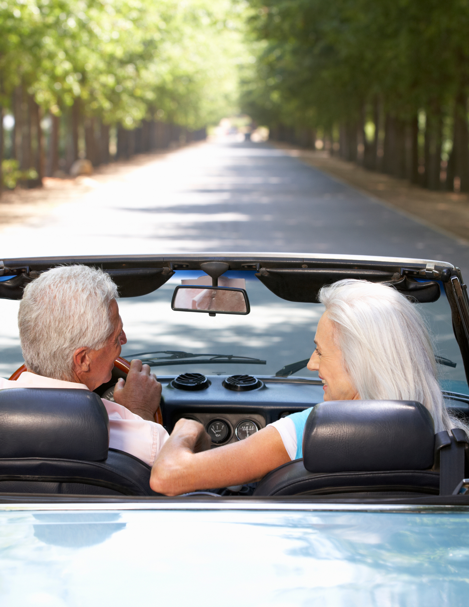 Older couple driving convertible on sunny tree-lined road